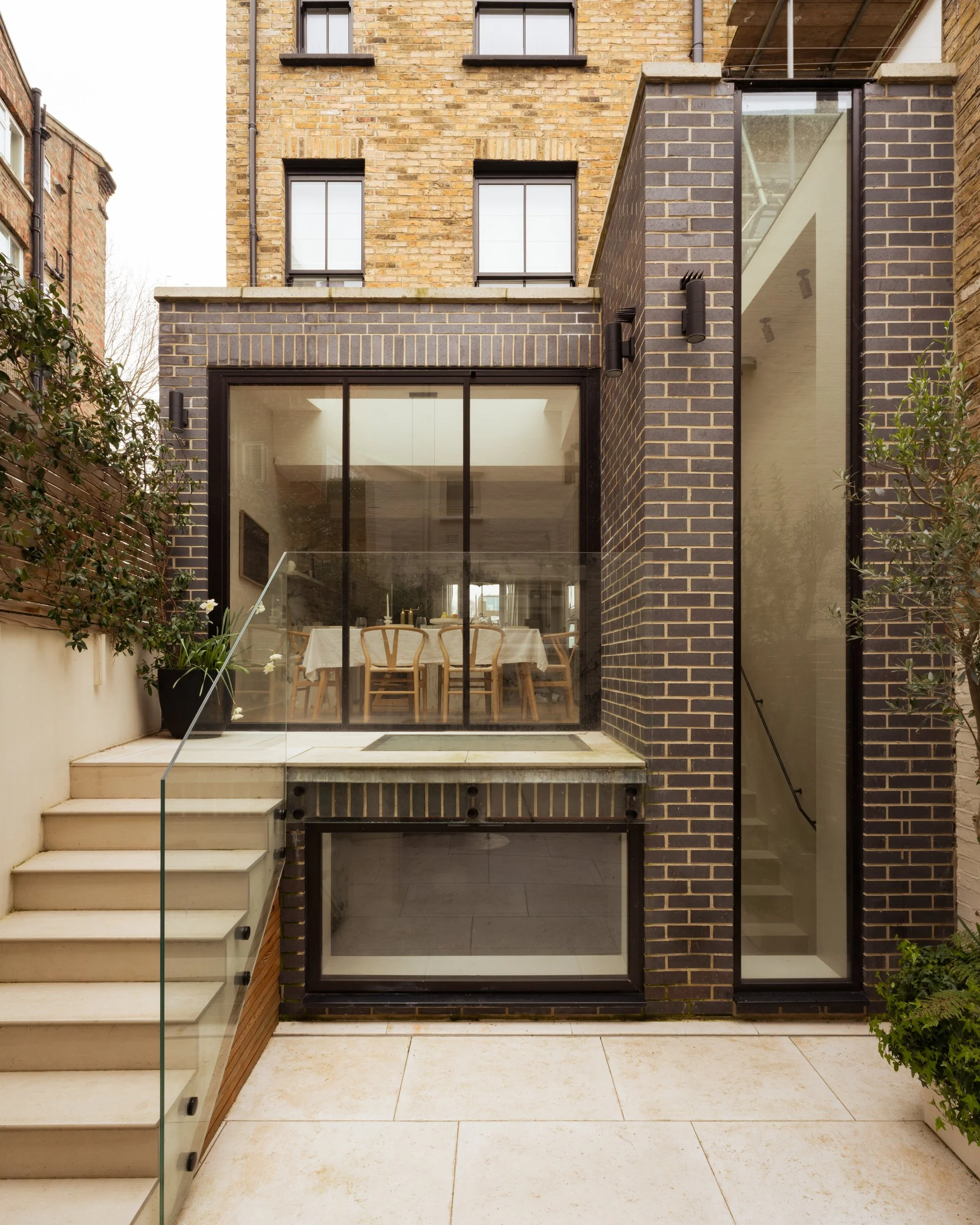 Modern brick building with large glass doors leading to an outdoor patio, staircase with glass railing, and potted plants.