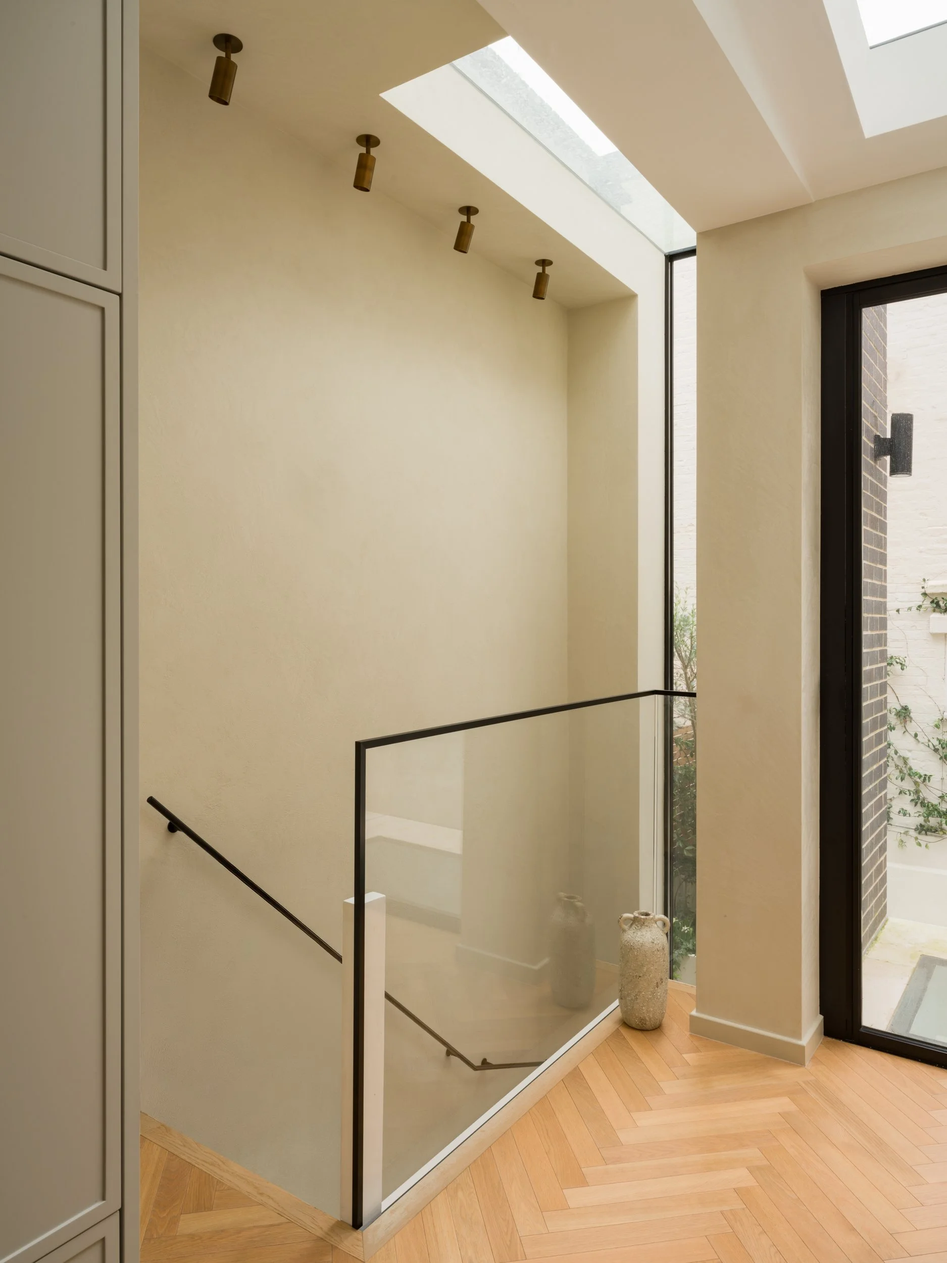 Interior view of a modern staircase with a glass railing, black handrail, and wooden flooring. Large windows and skylights provide natural light.