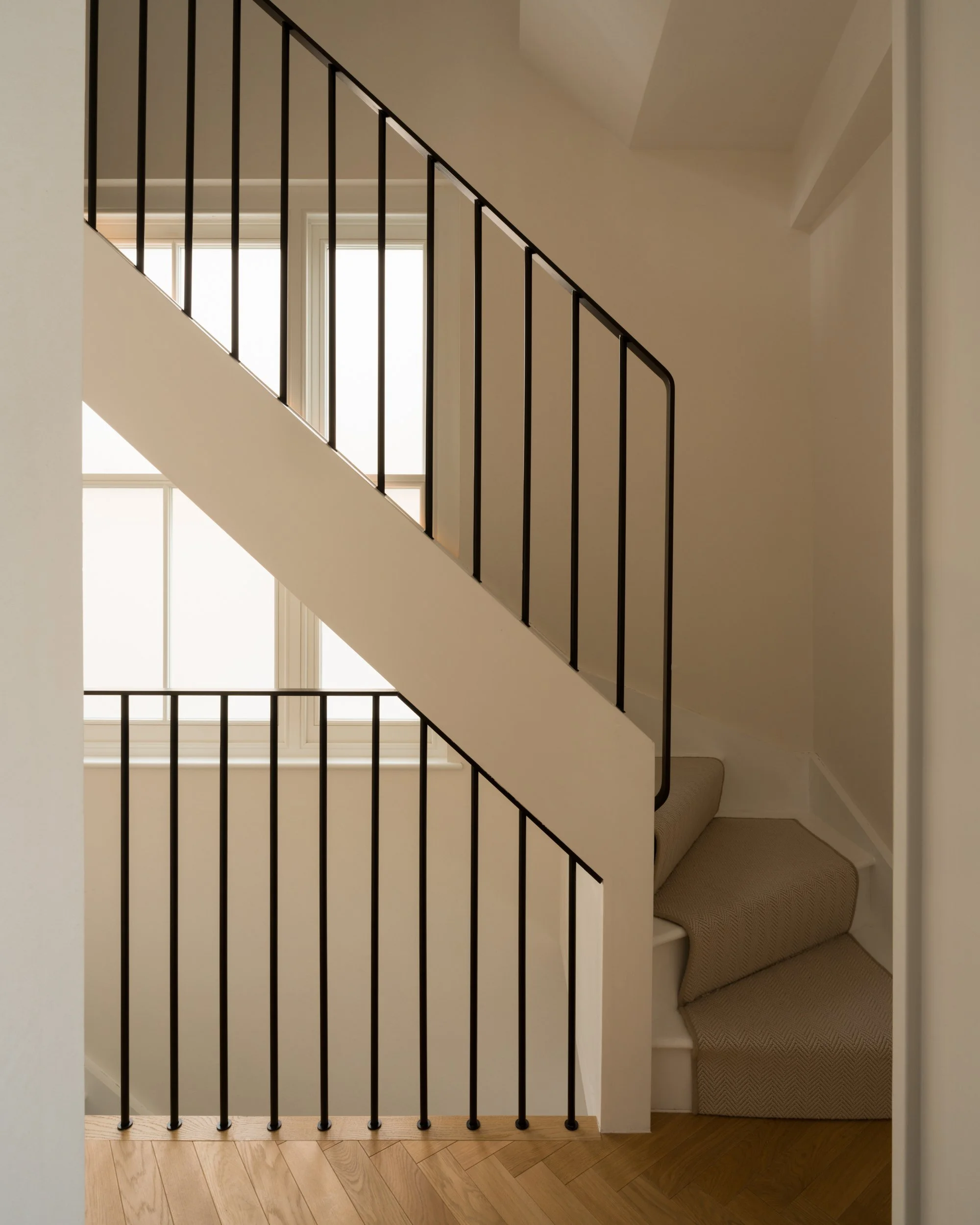 View of a staircase with black metal railing, a large window with multiple panes in the background, and a built-in window seat with beige cushions along the right side.