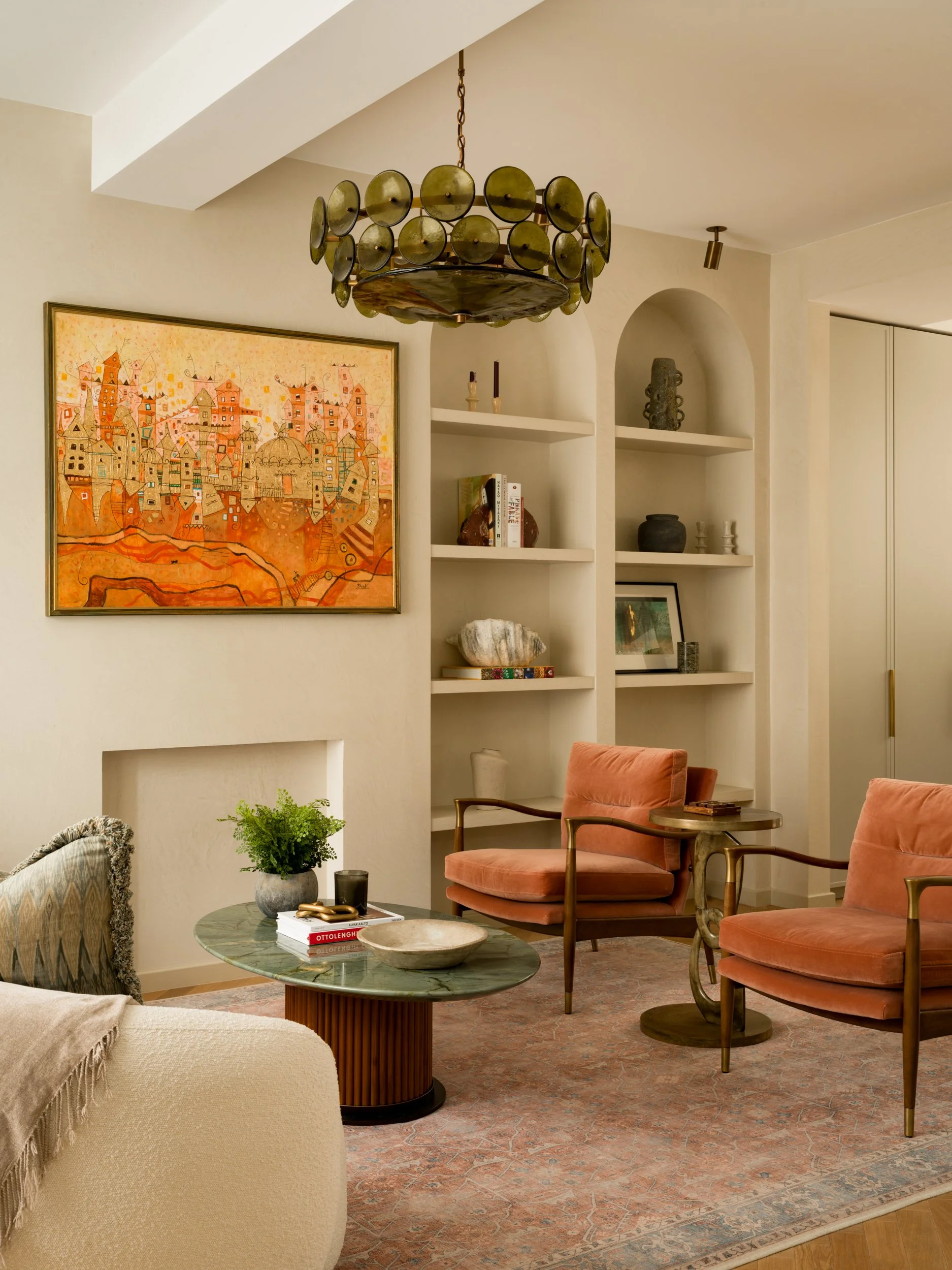 Living room with pink velvet armchairs, a marble coffee table, a colorful painting of a cityscape, built-in white shelves with decorative vases, a vintage chandelier, and a patterned rug.