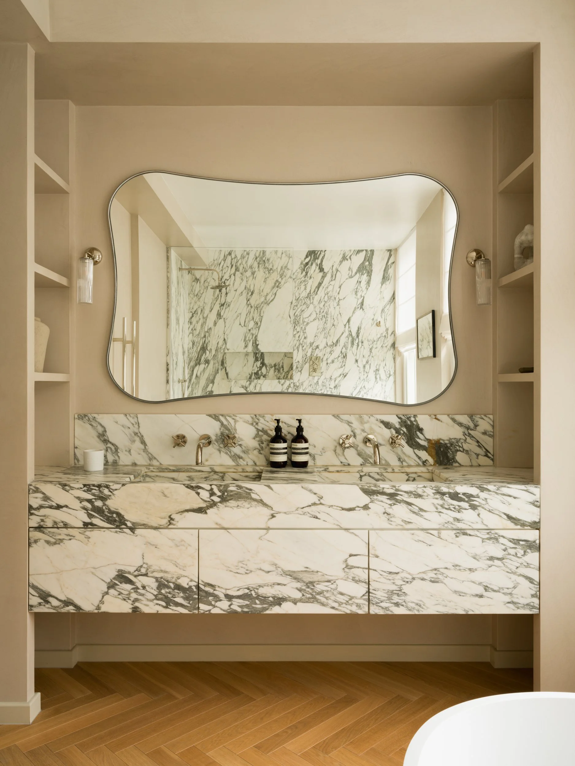 Modern bathroom vanity with marble countertop, large mirror, and two black bottles, wooden floor, and built-in shelving on sides.