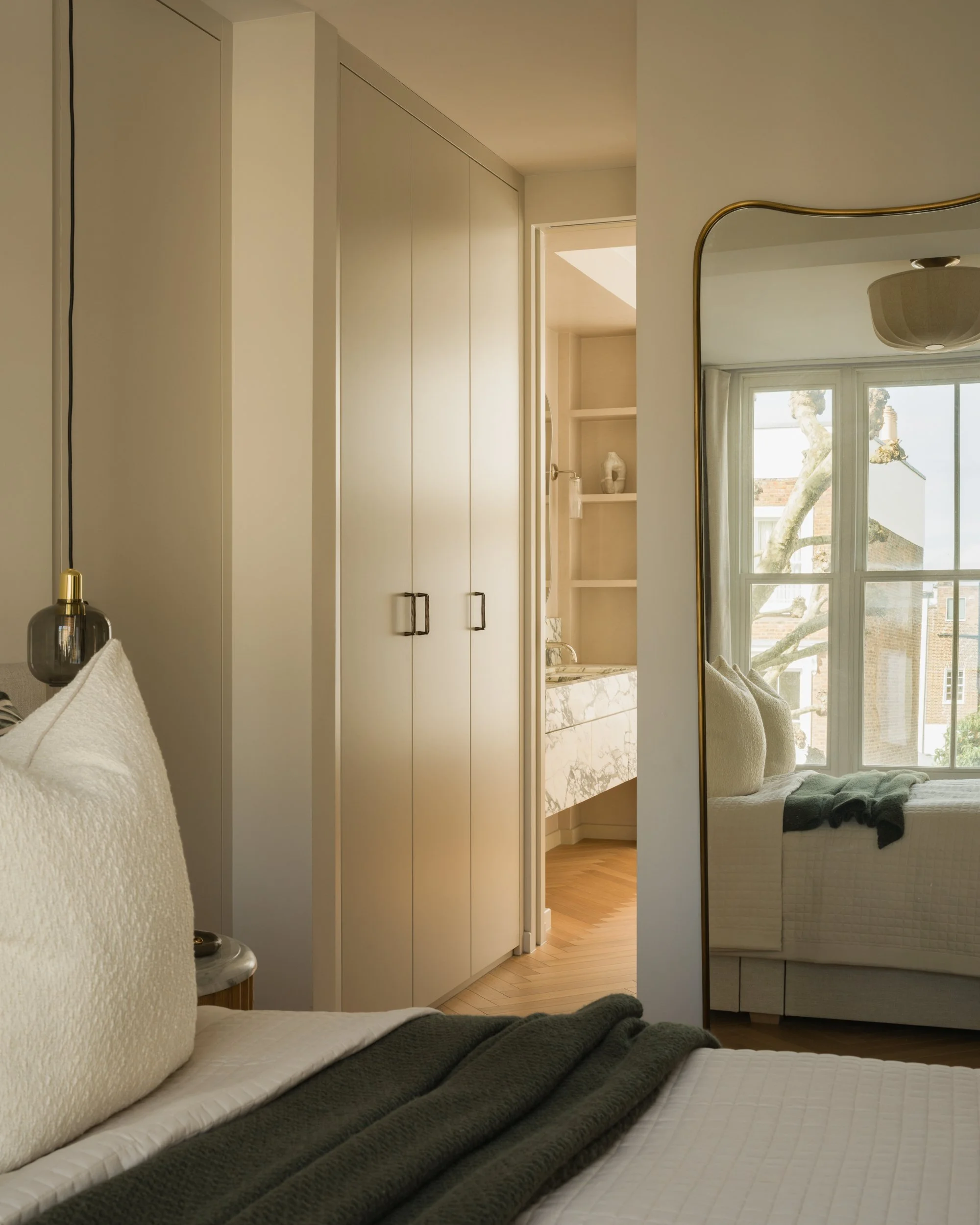 A cozy bedroom with a bed covered in a textured white bedspread and dark gray blanket, a cream-colored textured pillow, a small bedside table with a dark glass lamp, a built-in white wardrobe, a mirror reflecting a window with trees outside, and a glimpse into an adjoining room with marble countertops and a shelving unit.