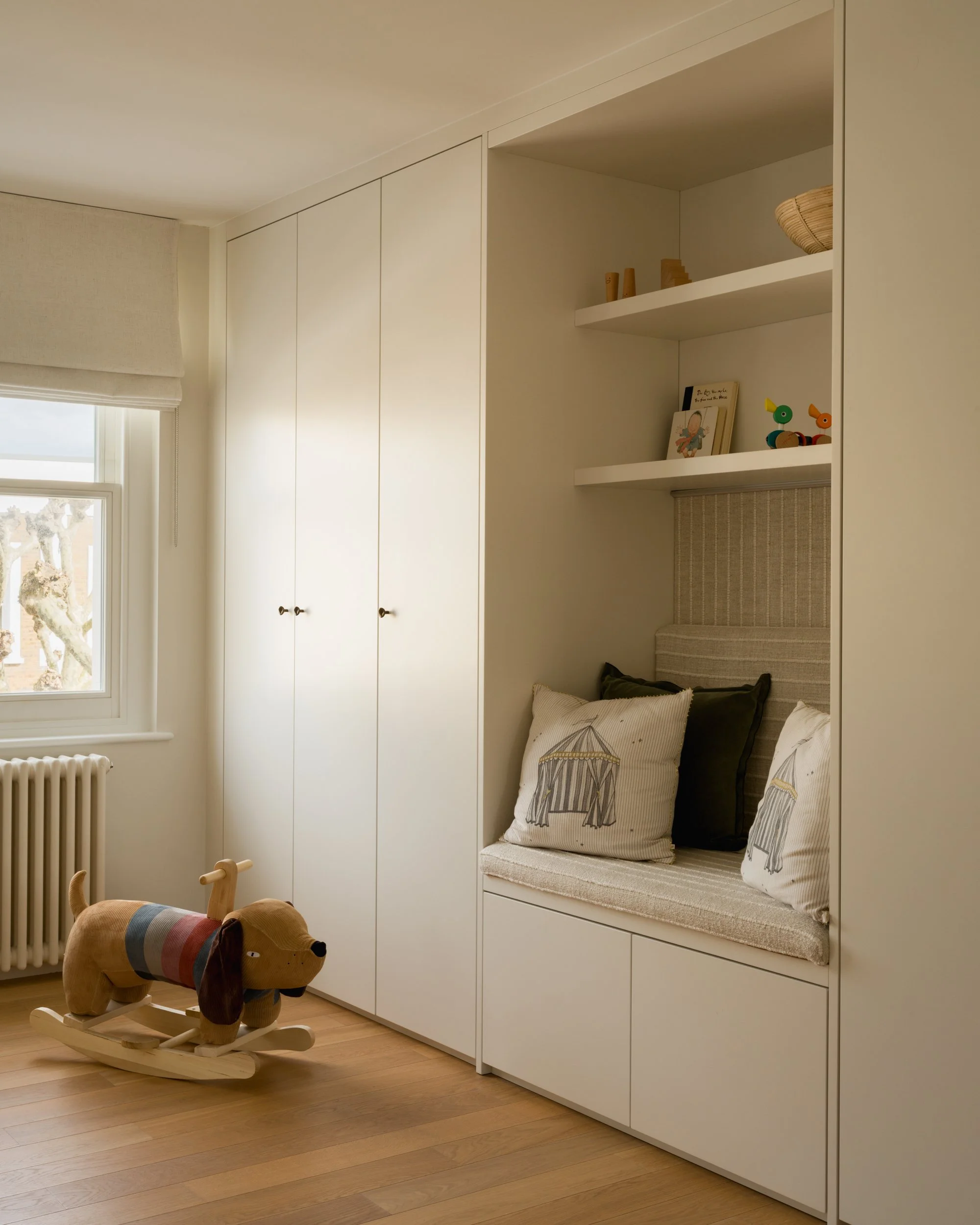 A cozy corner in a room with white built-in cabinetry, open shelving with decorative items and books, a window with a Roman shade, and a wooden rocking dog toy with a plaid blanket on its back on the wooden floor.