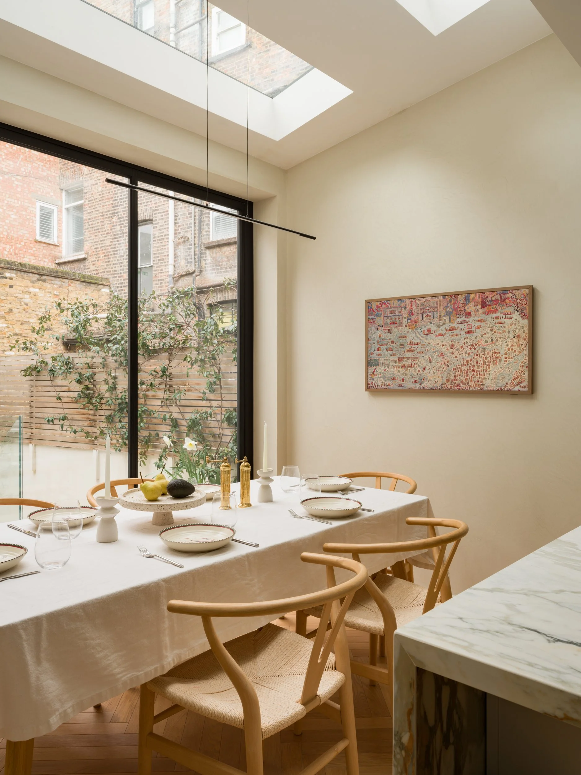 A dining room with a large window and a skylight, a round dining table with a white tablecloth, and wooden chairs. The table is set with plates, glasses, and cutlery. There is a decorative centerpiece with fruits and vases on the table. A painting hangs on the wall, and the exterior view shows brick buildings and a small garden.