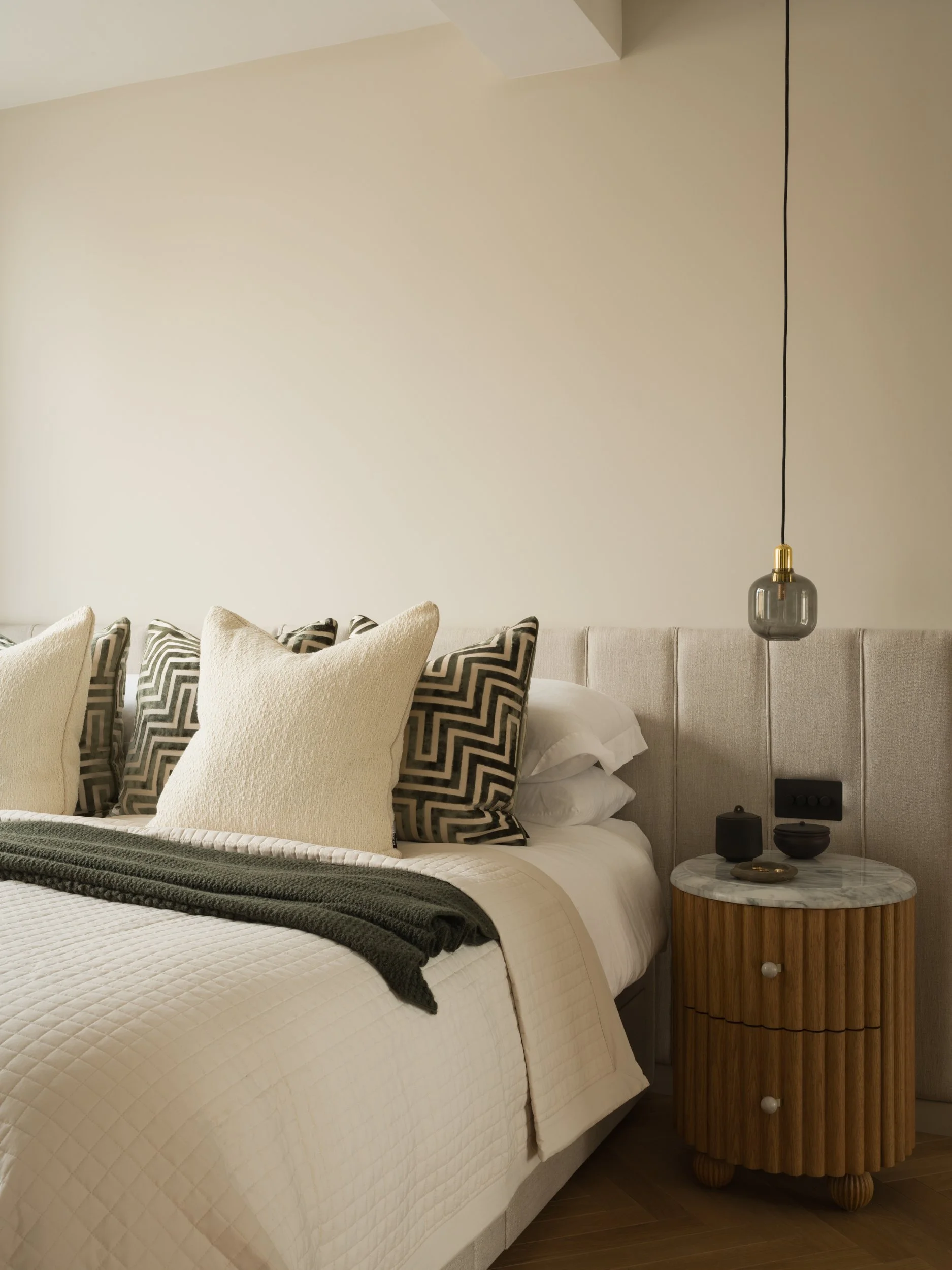 A neatly made bed with white quilt and multiple beige and black patterned pillows, a green blanket, and a small round wooden side table with a marble top holding decorative black and gold objects, against a beige upholstered headboard and plain beige wall with a black pendant light.