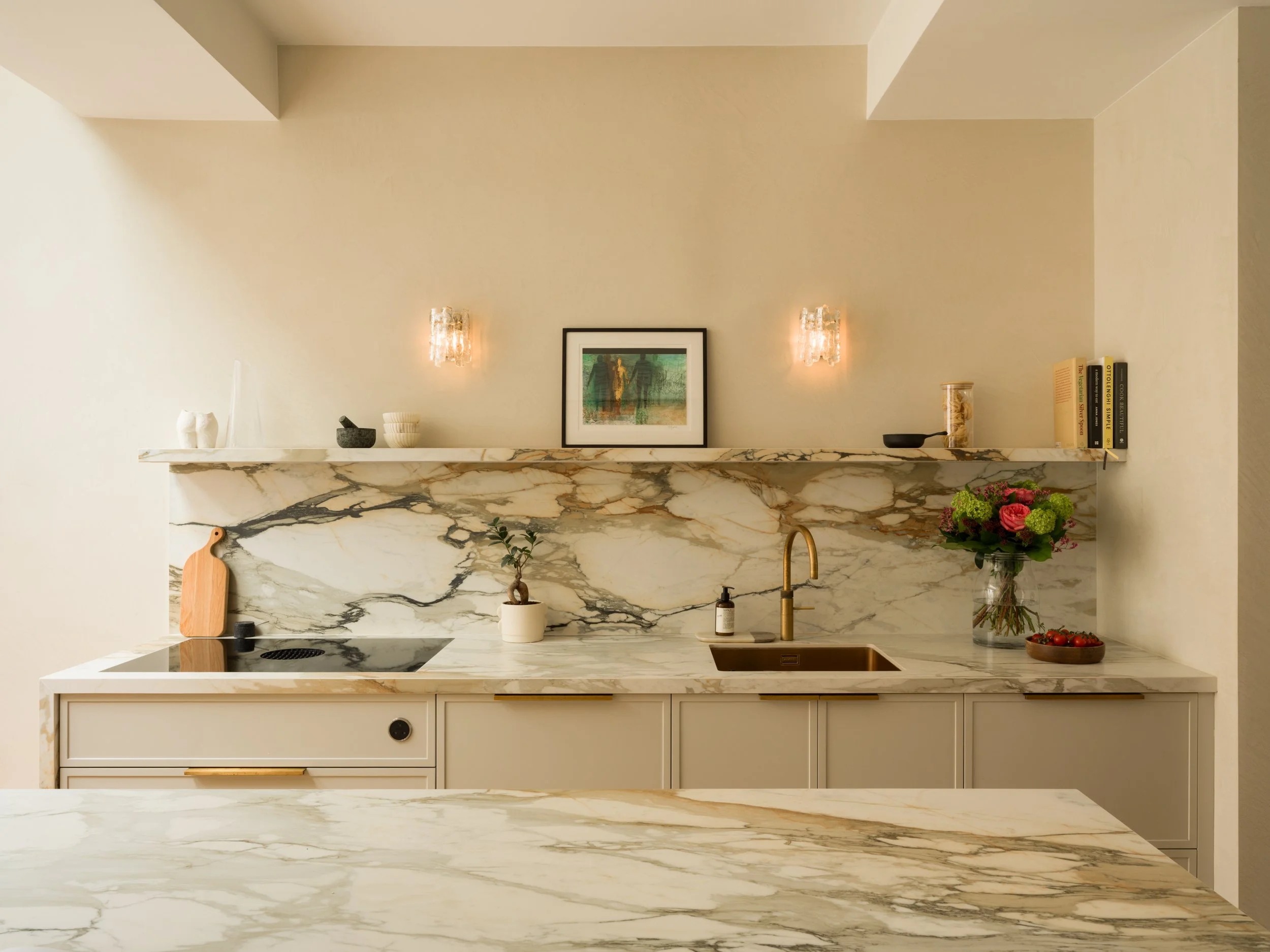 A modern kitchen with a marble countertop and backsplash, beige walls, framed artwork, small decorative items, and fresh flowers on the counter.