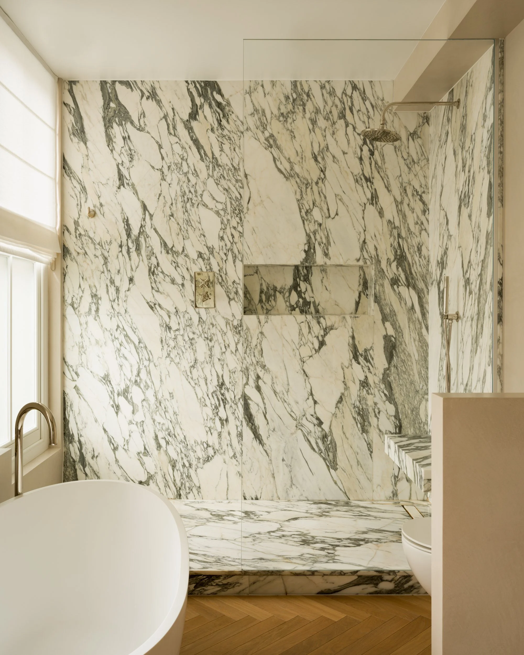 A bathroom with white and gray marble walls and flooring, a walk-in shower with a rain showerhead, a window with a white blind, and wooden flooring outside the shower.