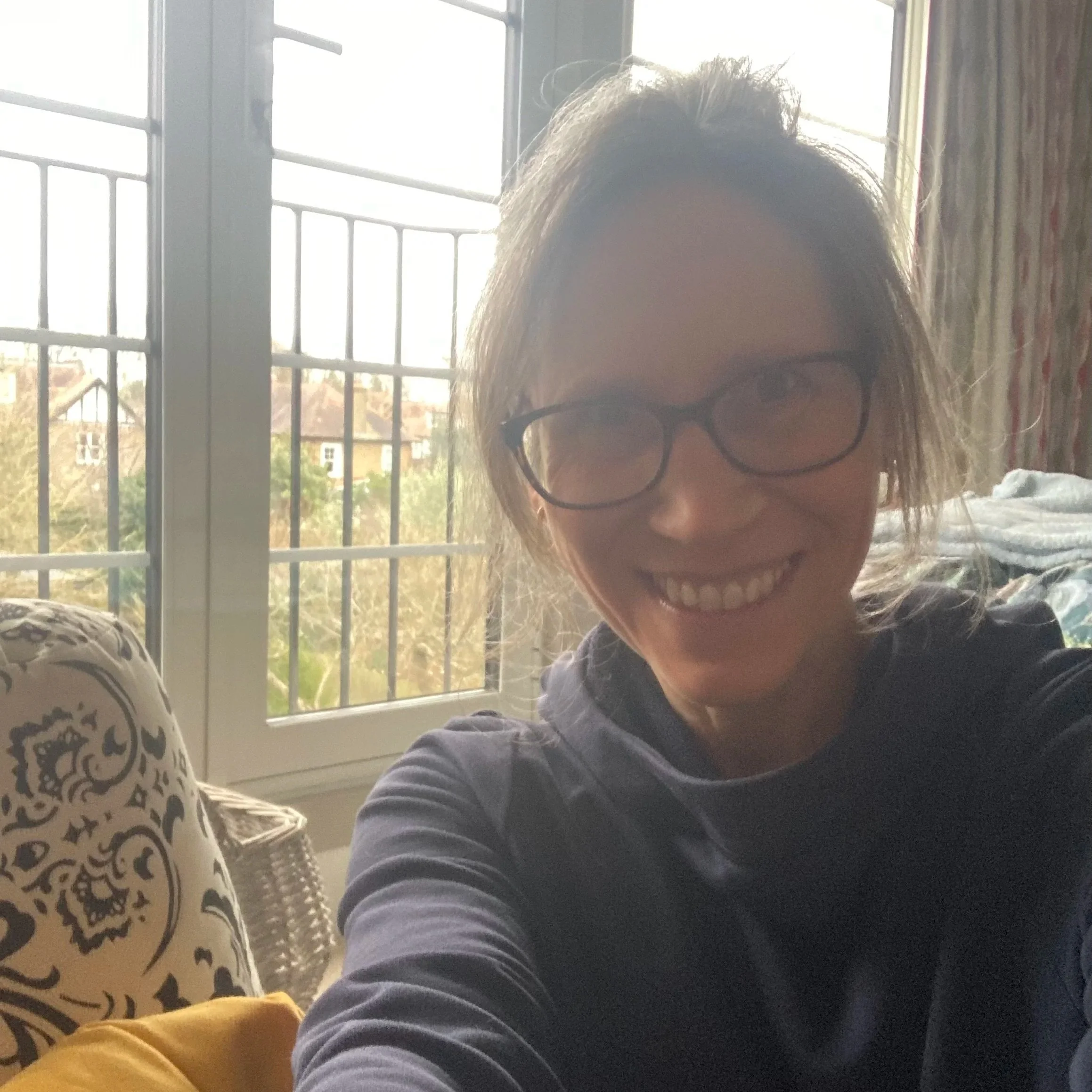 Caroline sitting on her bedroom floor next to a white and black patterned yoga bolster, wearing a dark blue hoodie and glasses. In the background is a view of houses and gardens out of the window and the edge of lots of soft furnishings.