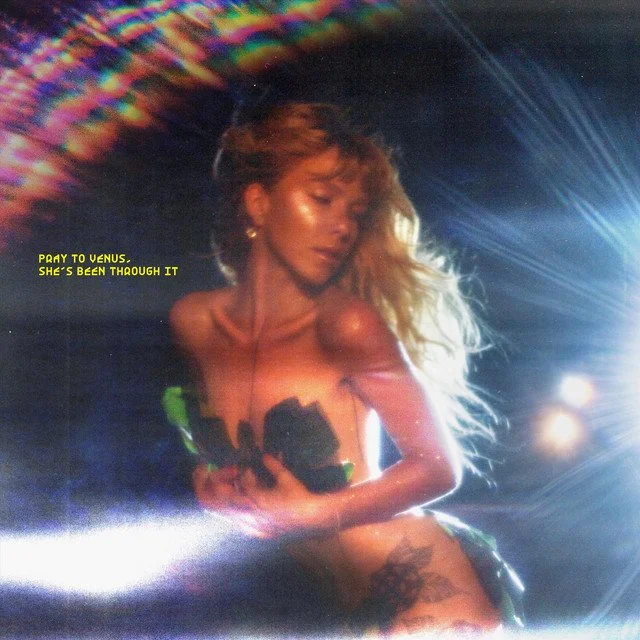 A woman with long hair dancing or performing on stage under colorful lights, with a bright spotlight shining from the right. There is a yellow caption that reads "Pay to Venus, she's been through it."
