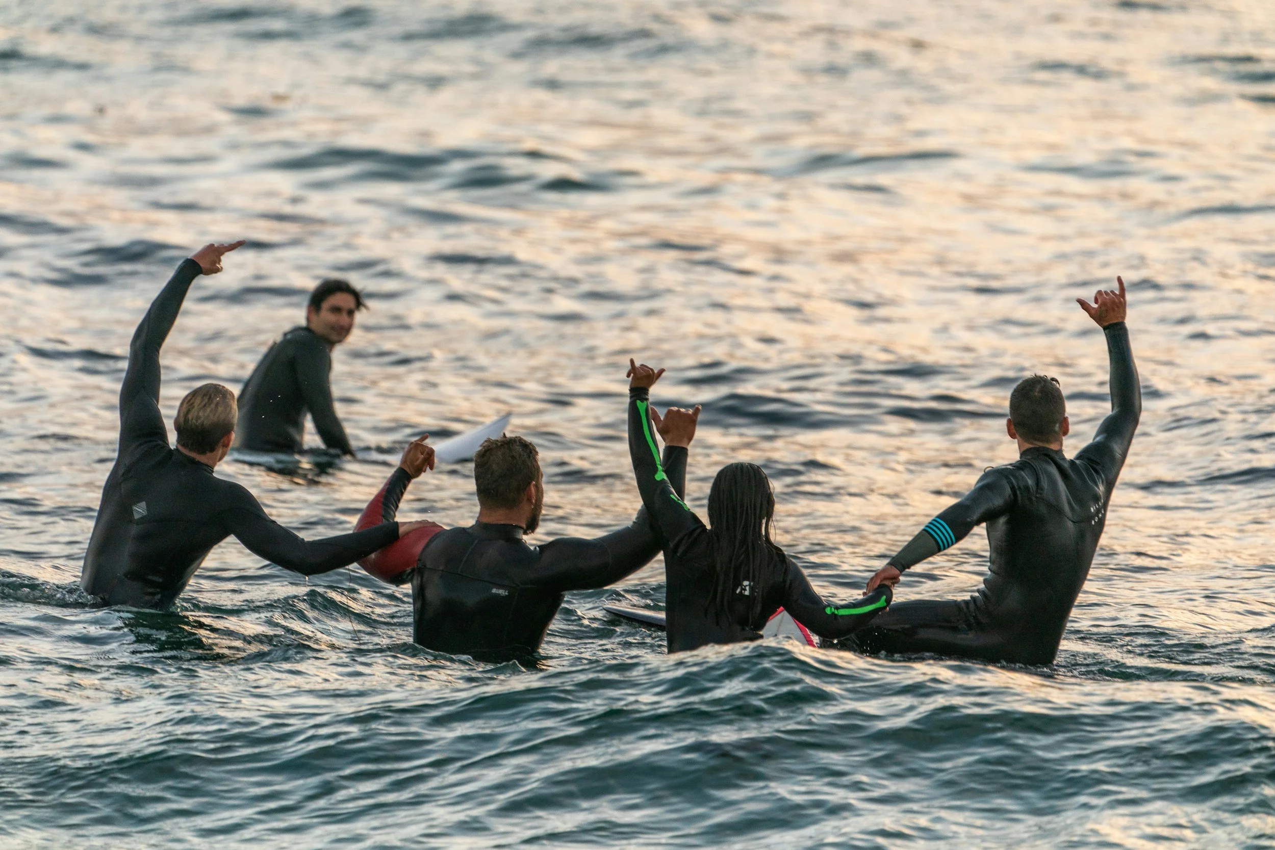 A group of five people in wetsuits holding surfboards and making celebratory gestures while in the ocean at sunset.