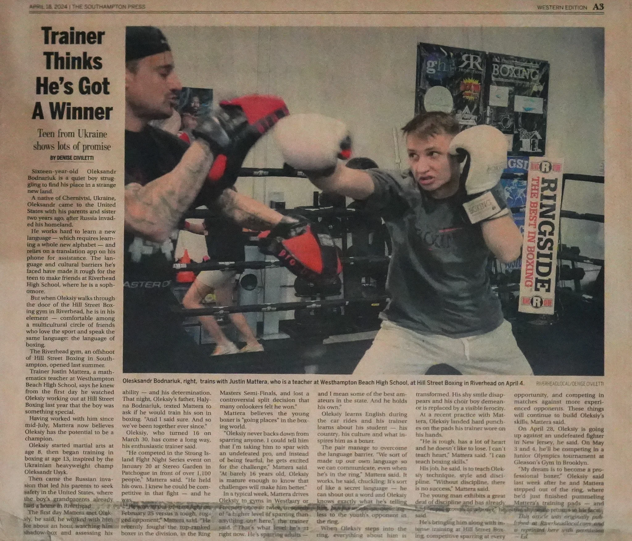 A young boy wearing boxing gloves and gray shirt sparring with a man in black and red boxing gloves at Westhampton Beach High School gym, with boxing equipment and posters in the background.