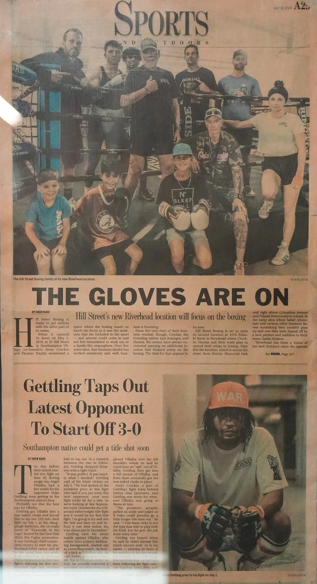 A newspaper page featuring a photo of a group of people, including children and adults, in a boxing gym. The headline reads "The Gloves Are On," with articles about a new boxing location and a boxing match. The photo shows the group posing in a boxing ring, some wearing boxing gloves and sports attire, with a young girl dressed as a boxer sitting in front.