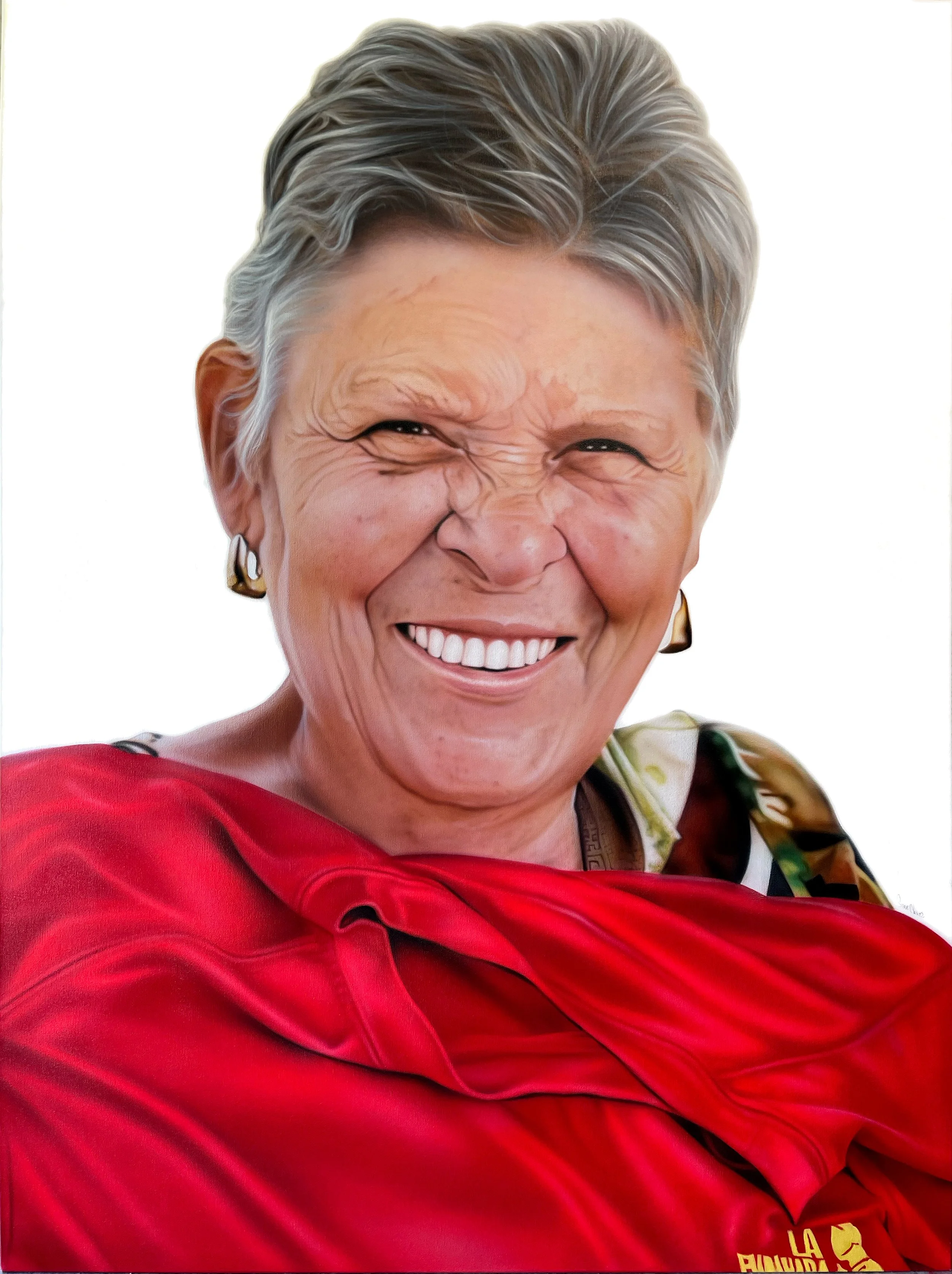 A smiling elderly woman with short gray hair, wearing gold earrings and a red jacket, is crossing her arms against a plain white background.