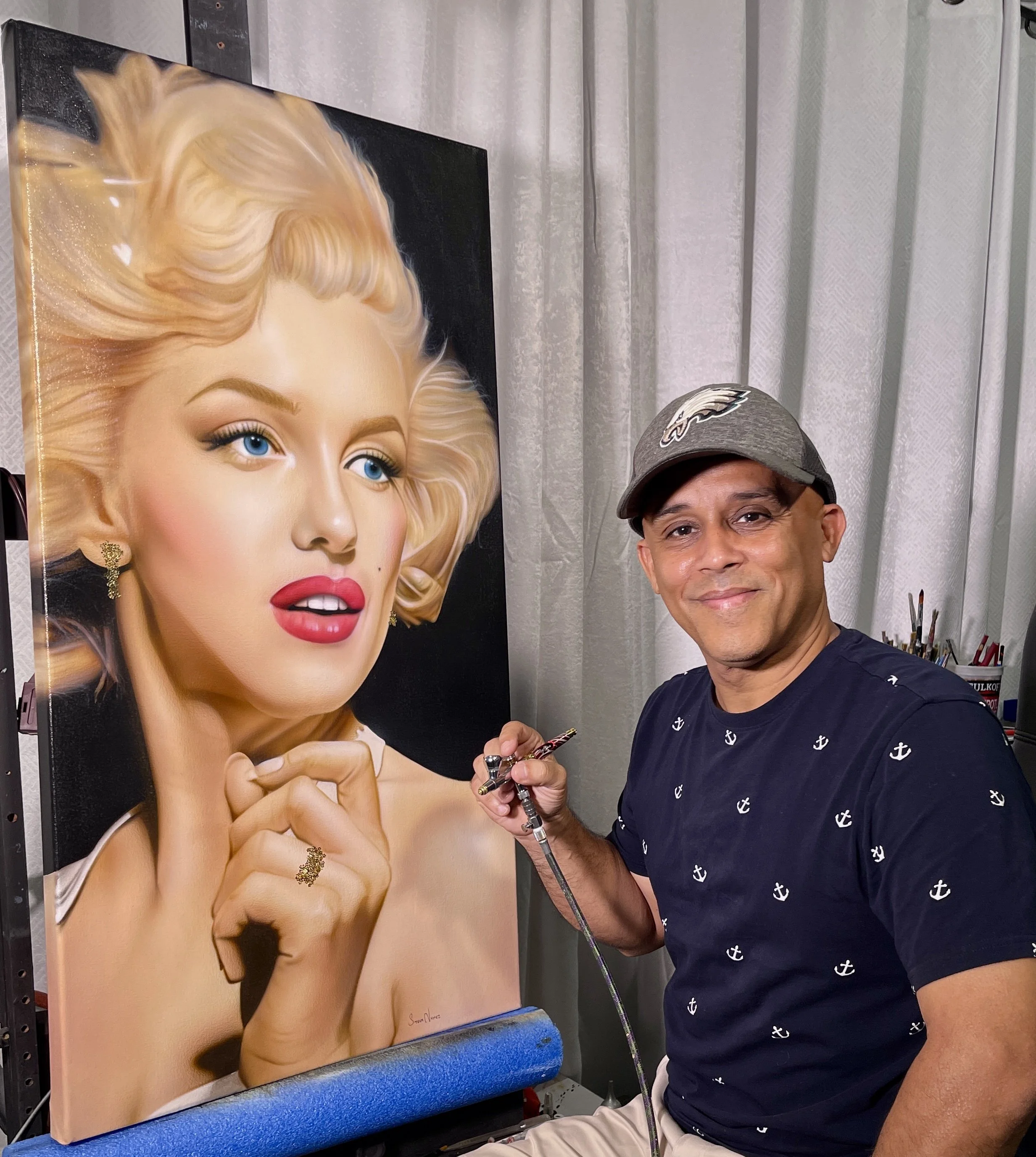 A man in a navy blue shirt with white anchor patterns and a gray cap standing next to a large portrait of a woman with blonde hair and bright makeup. The man is holding a tool, possibly an airbrush, and smiling at the camera. They are in an indoor setting with curtains in the background.
