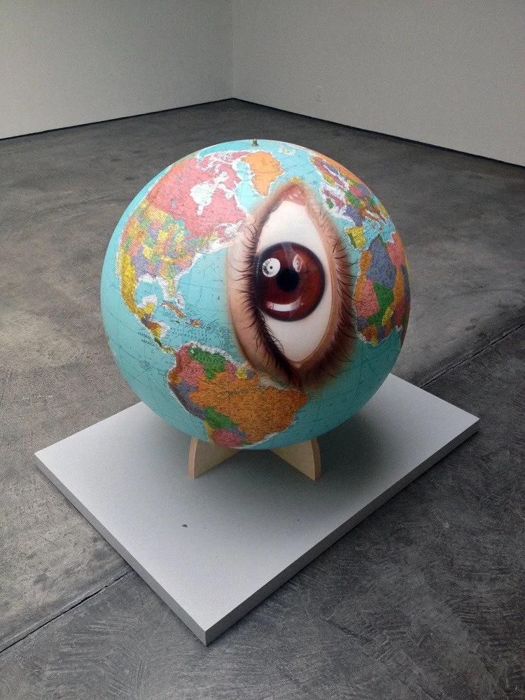 A globe with a large realistic human eye in the center, displayed on a white stand in an art gallery.