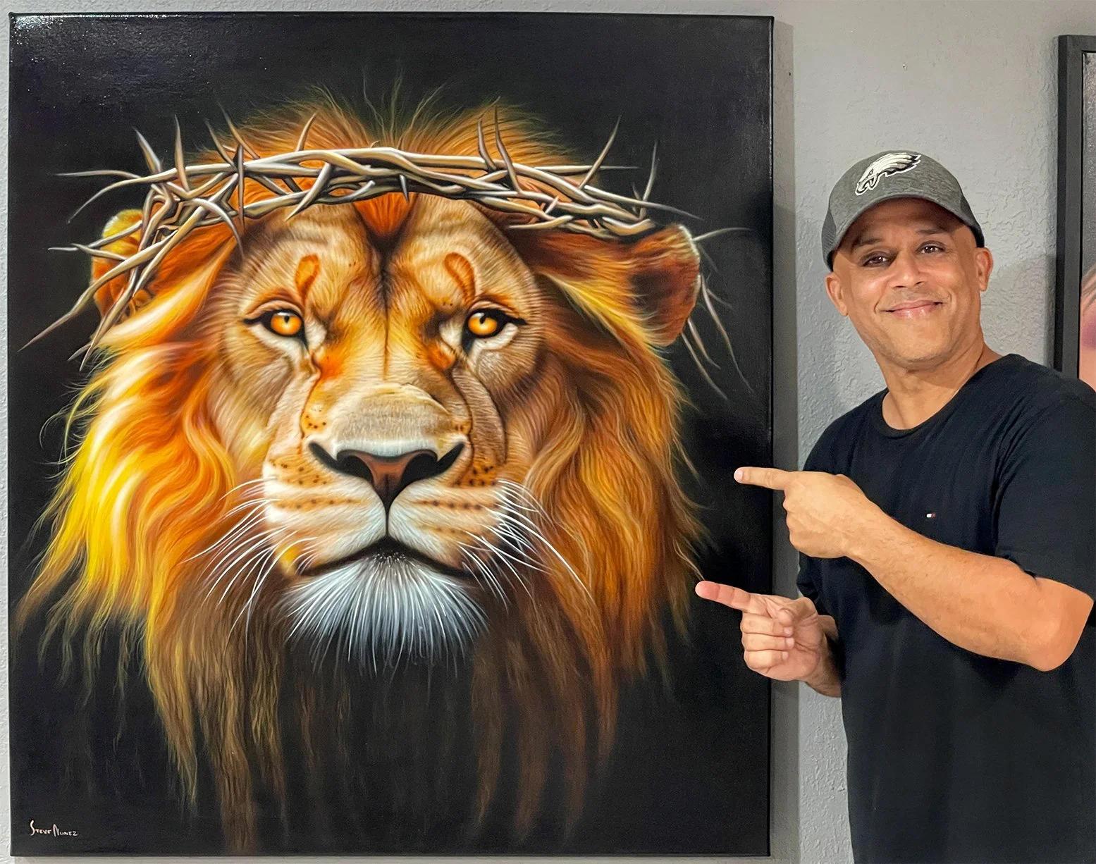 A man in a gray cap and black T-shirt pointing at a large, colorful painting of a lion wearing a crown of thorns. The painting depicts the lion with detailed orange and yellow fur against a black background.