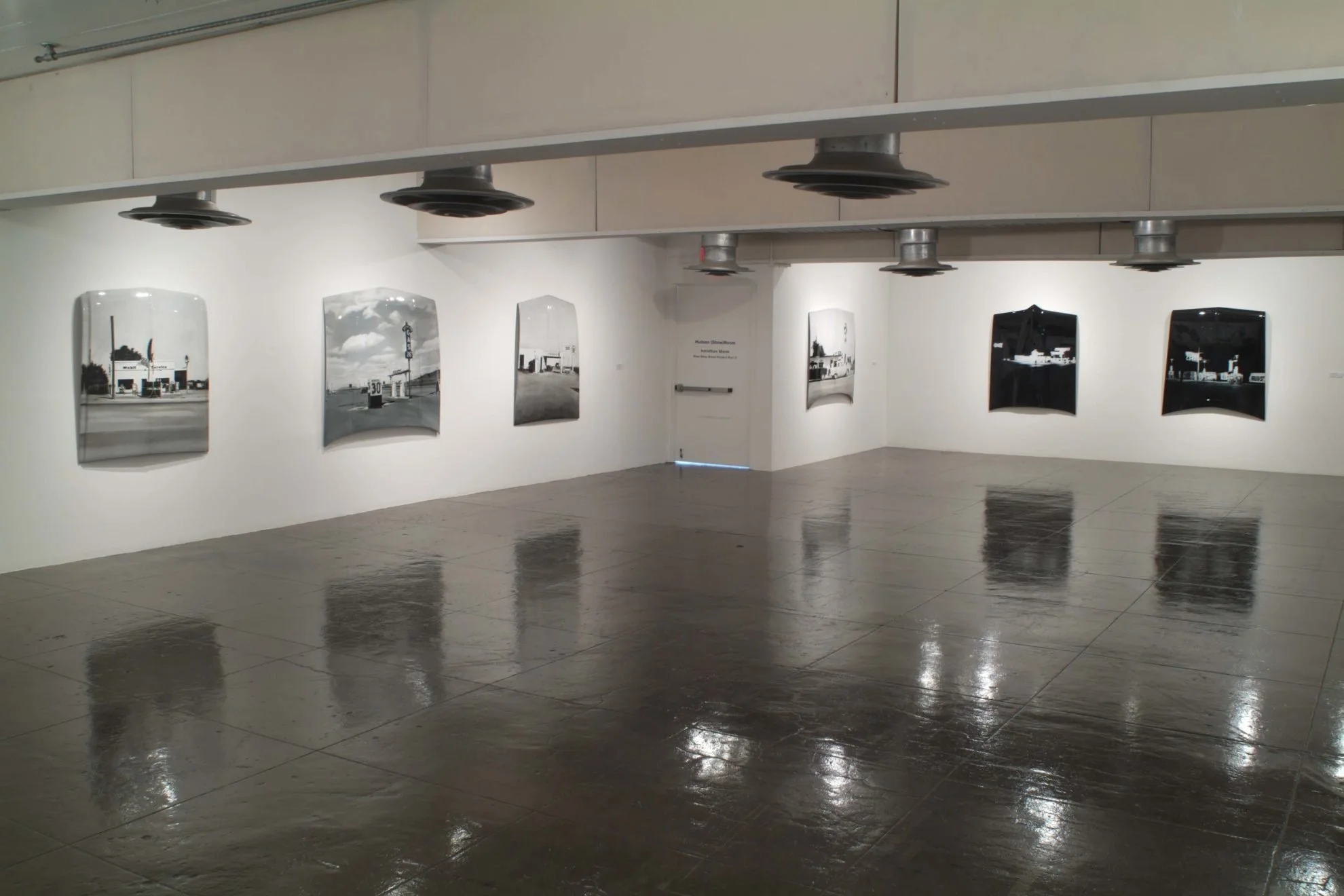 Art gallery with black and white photographs of buildings and street scenes on white walls, polished concrete floor, overhead industrial lighting.
