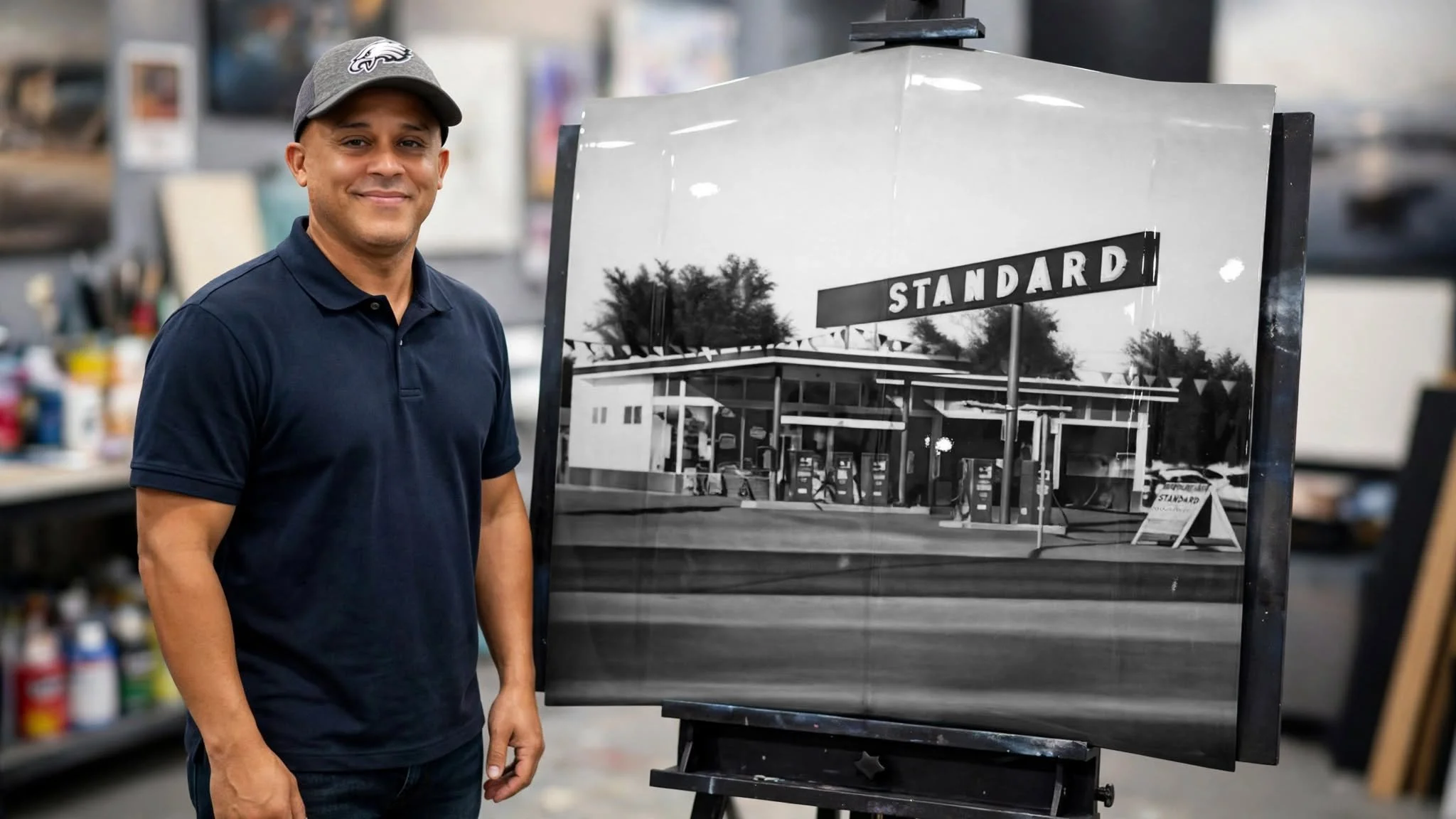 A man wearing a gray cap and a navy polo shirt standing next to a large black-and-white photograph of a gas station with a prominent sign reading 'STANDARD' indoors.