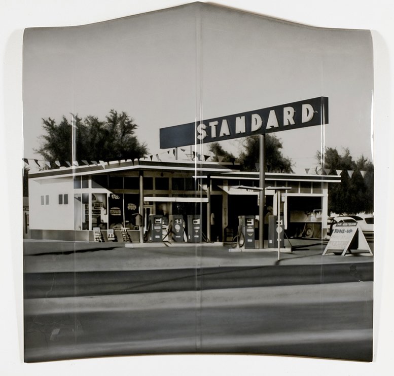 A vintage black-and-white photo of a gas station with a large sign that reads "STANDARD" on top, several gas pumps outside, and a small building, possibly a convenience store, in the background.