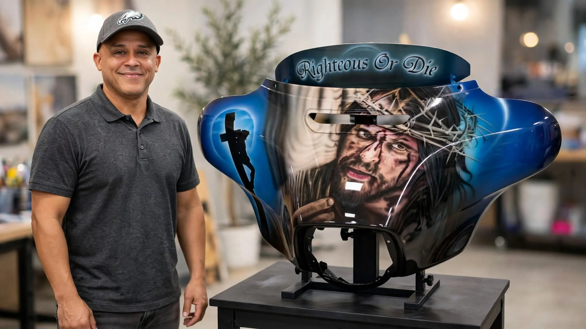 A man wearing a gray polo shirt and a gray cap with a logo, standing next to a motorcycle helmet with detailed art, inside a workshop or studio.