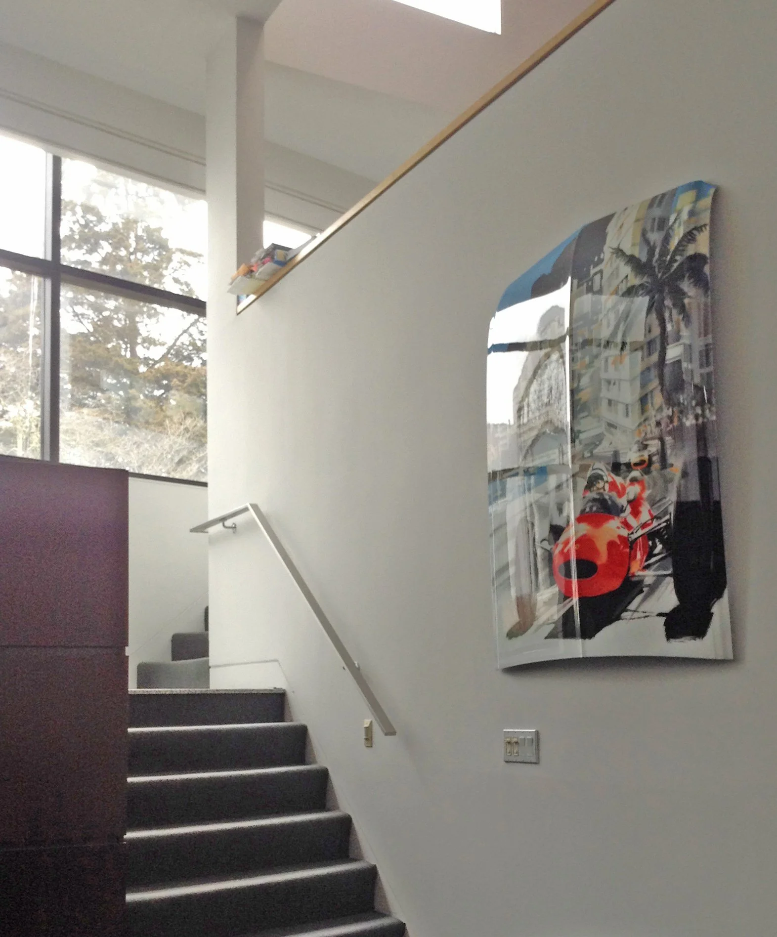 Interior view of a modern staircase with black steps, white walls, and a glass railing. Large windows let in natural light, revealing trees outside. A colorful abstract painting hangs on the wall.