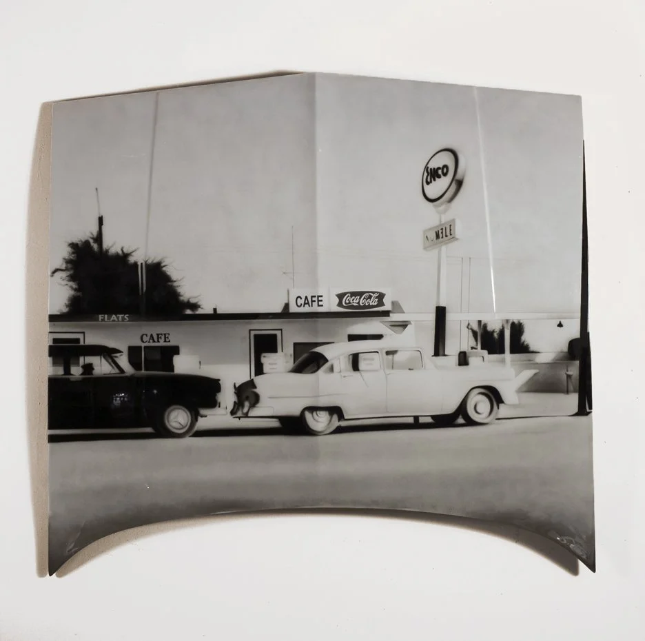 Black and white photo of a vintage roadside scene with cars parked in front of a cafe, featuring signs for Coca-Cola and Anco, with a large sign displaying the name "N. Malle".