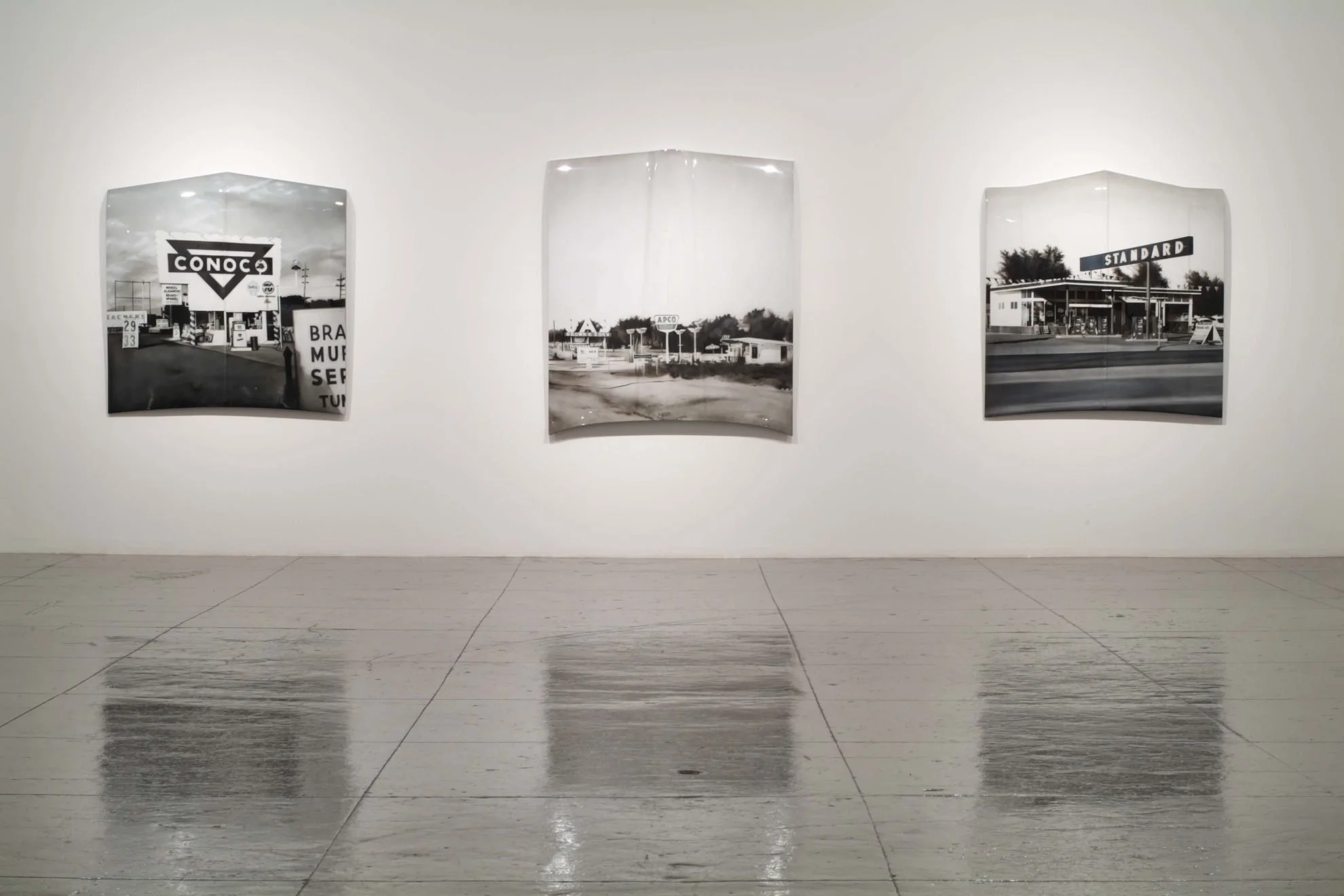 Black and white photographs of vintage gas stations displayed on a white gallery wall.
