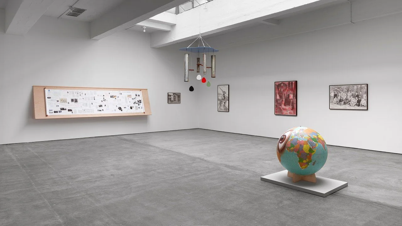 An art gallery with white walls, concrete floors, and artwork including framed photographs, paintings, a globe sculpture with a map, and a hanging mobile.