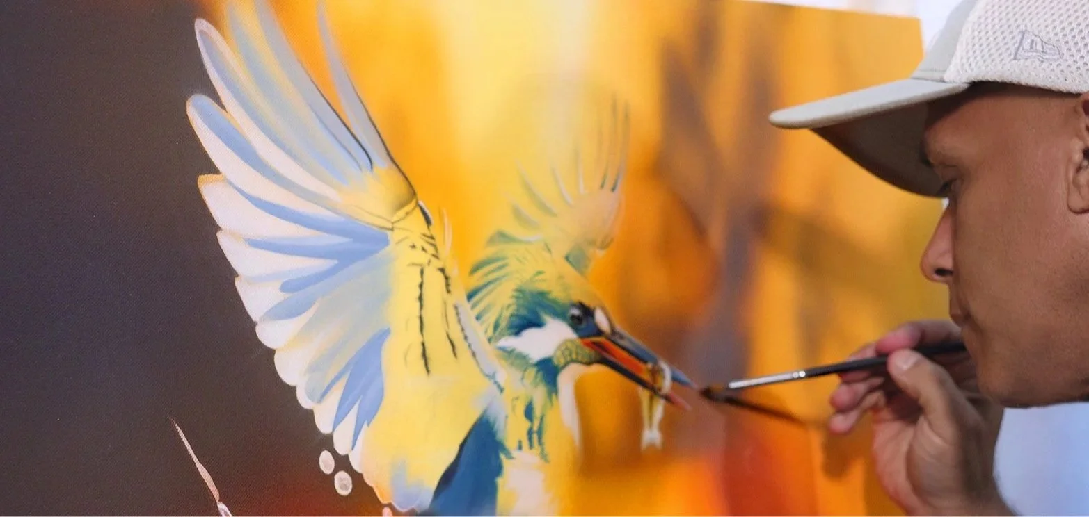 An artist painting a colorful, detailed image of a yellow and blue bird, possibly a parrot, on a canvas.