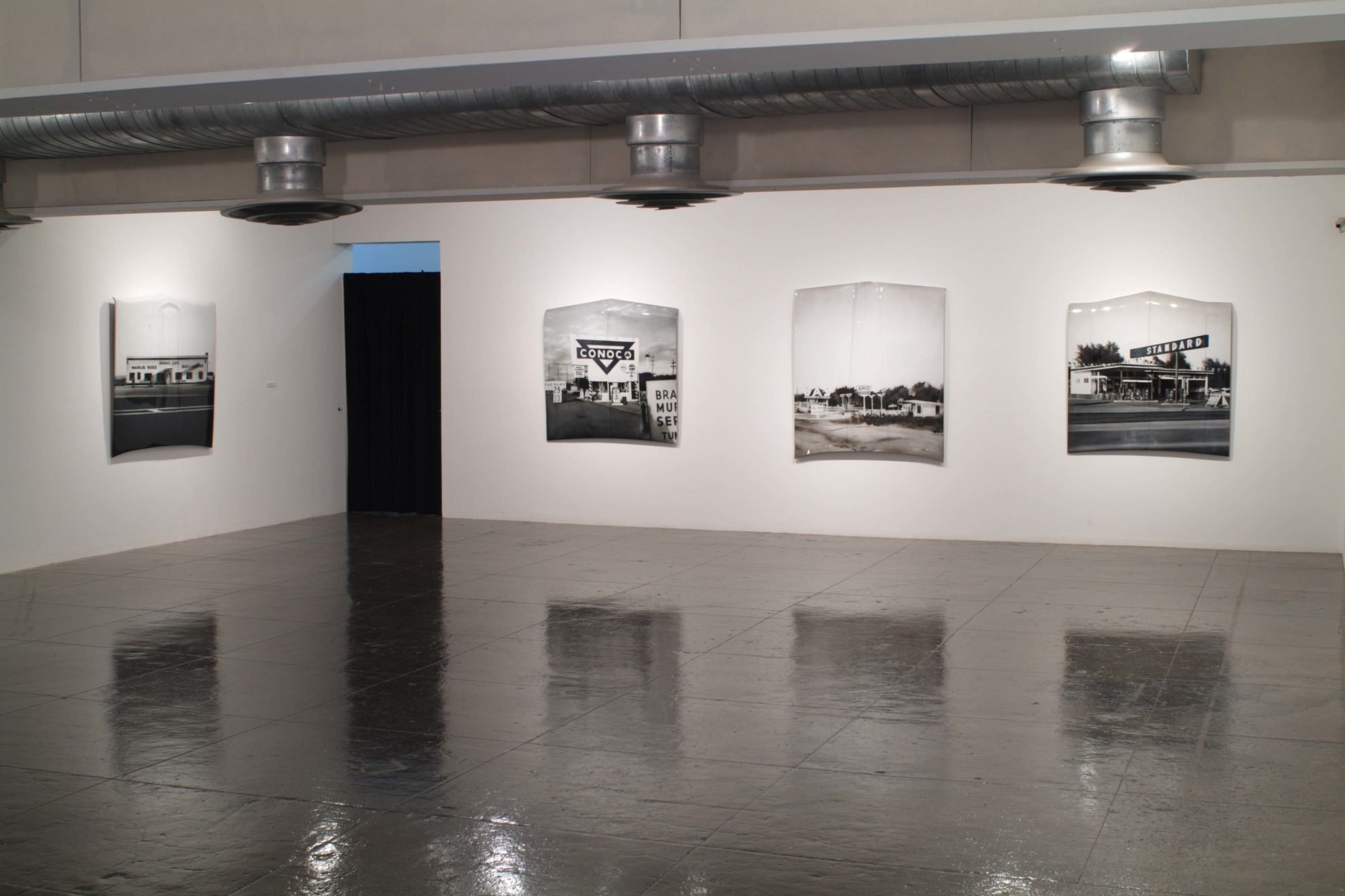 An art gallery with black-and-white photographs of a gas station, road signs, and a building displayed on white walls.