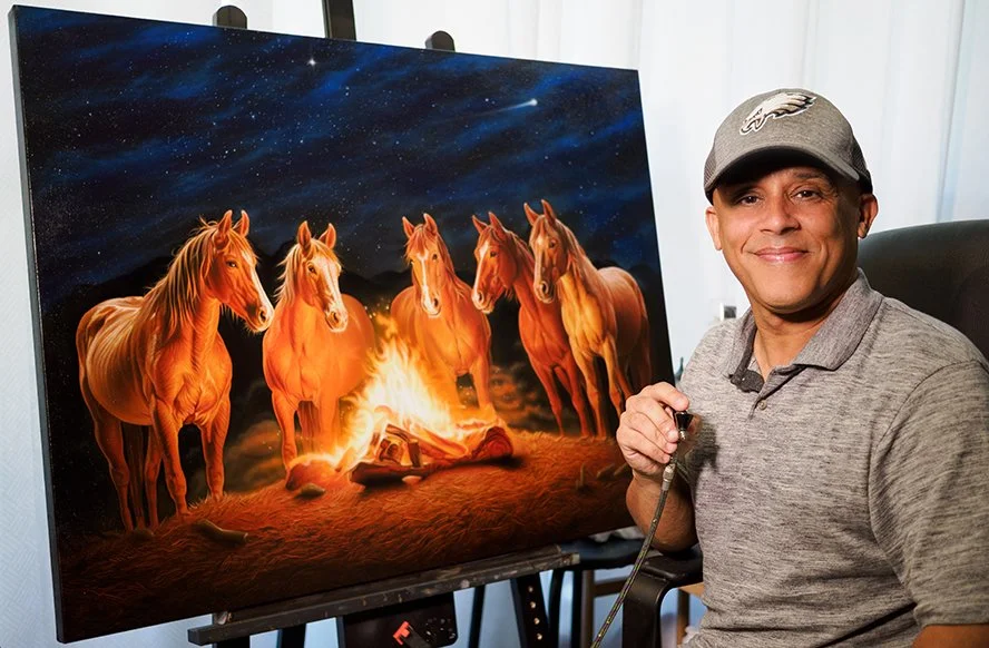 A man in a gray shirt and Philadelphia Eagles cap smiling, holding a paintbrush, sitting next to a painting of five horses around a campfire under a starry night sky.