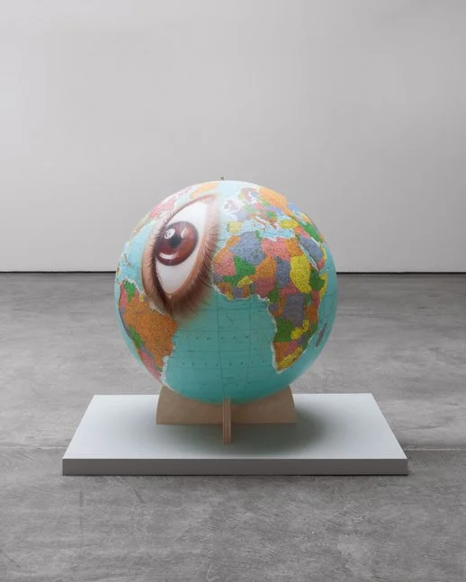 A globe with a painting of a human eye on its side, placed on a white platform in a gallery setting.