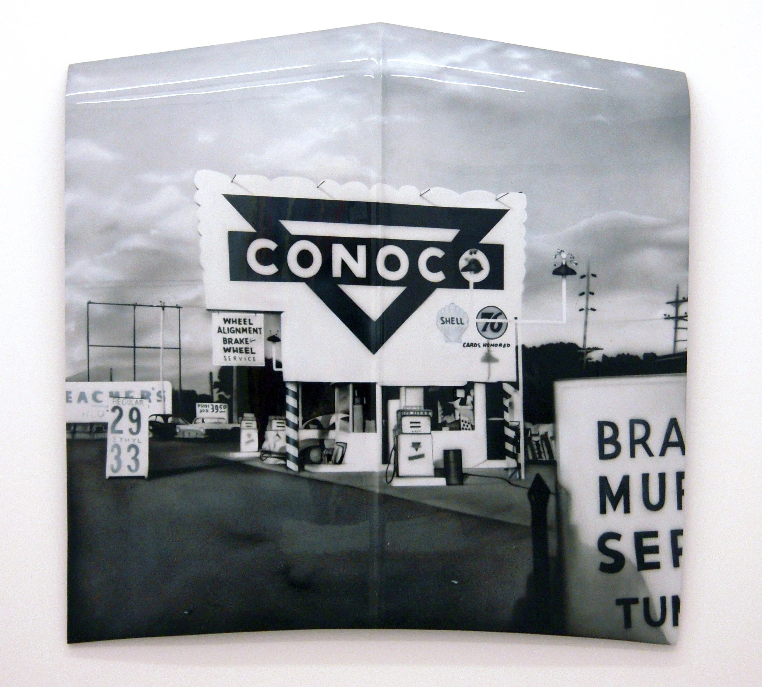 A black and white photograph of a street scene featuring various road signs, including a Conoco gas station sign, speed limit signs, and other traffic notices, with cars parked in the background.