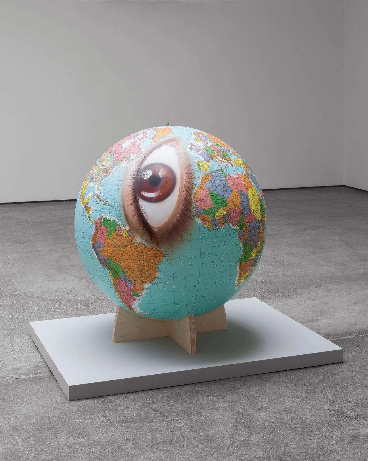 An art installation of a globe with a large realistic human eye painted on it, positioned on a wooden stand on a white platform in a gallery with gray floors and white walls.