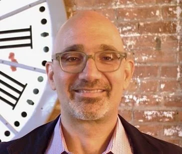 A man with glasses and a beard smiling in front of a large clock and a brick wall.