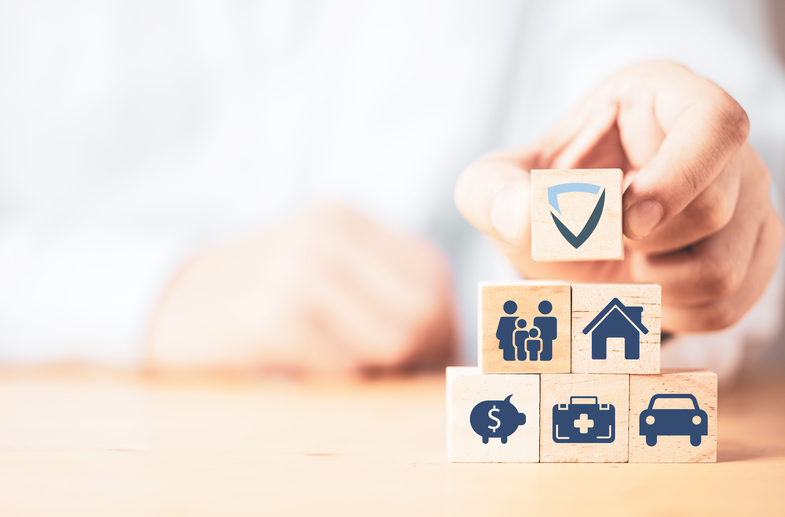 Wooden blocks with symbols representing family, home, savings, health, transportation, and a shield for protection, arranged in a pyramid formation.