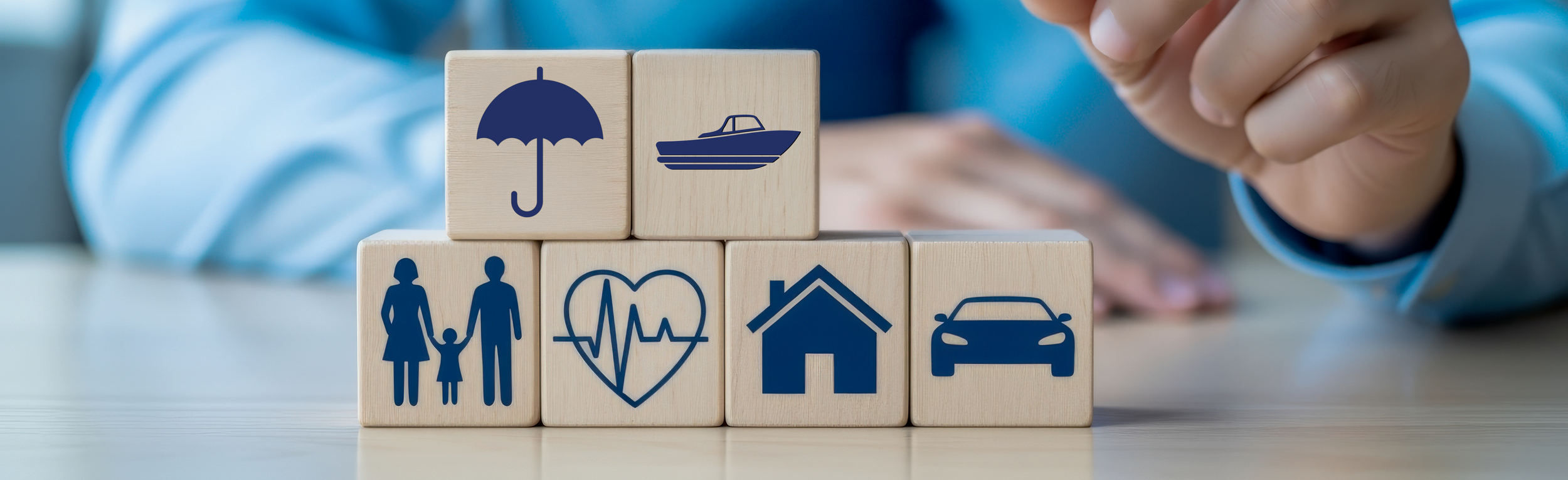 Stacked wooden blocks with blue icons representing umbrella, boat, family, heart health, house, and car, arranged on a table.