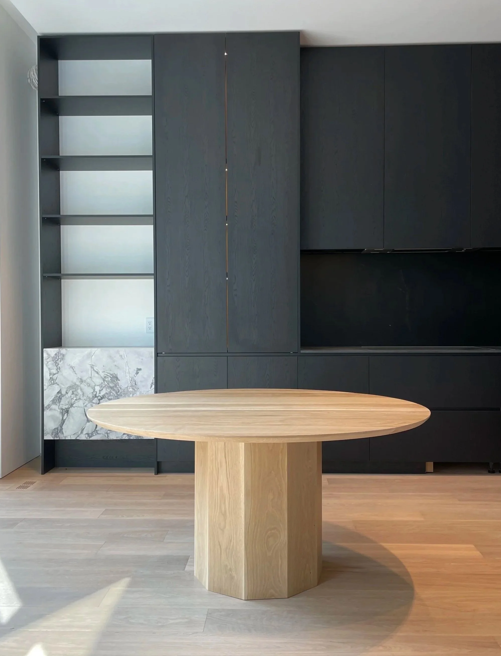 Modern kitchen with dark cabinets, light wood table, marble backsplash, and light-colored wooden flooring.