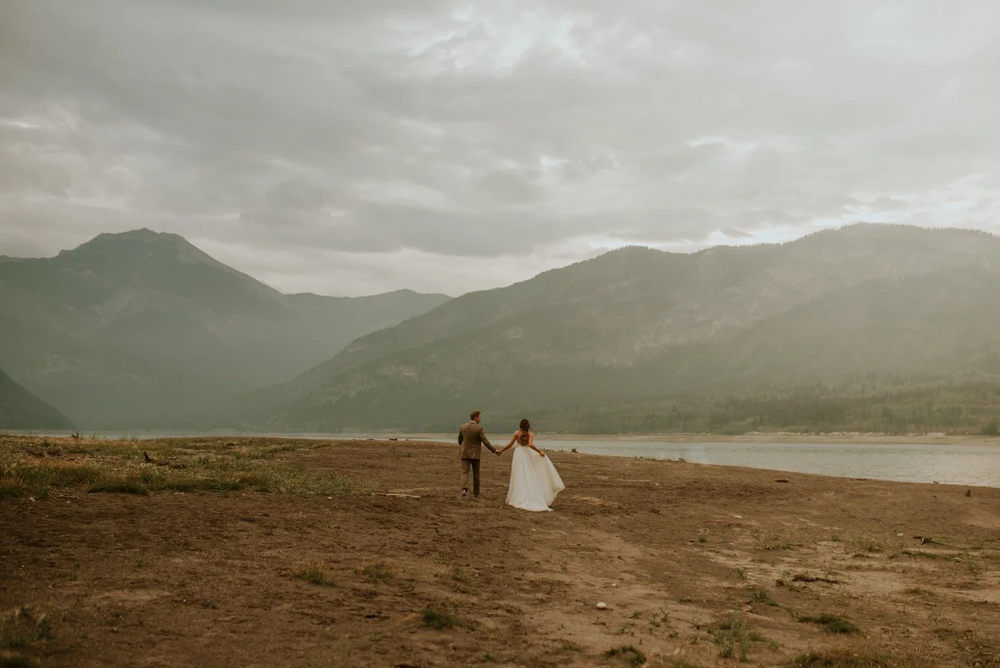 A bride and groom holding hands walking on a rugged terrain near a river with mountains in the background and overcast sky. Elopement in Barrier Lake Kananaskis