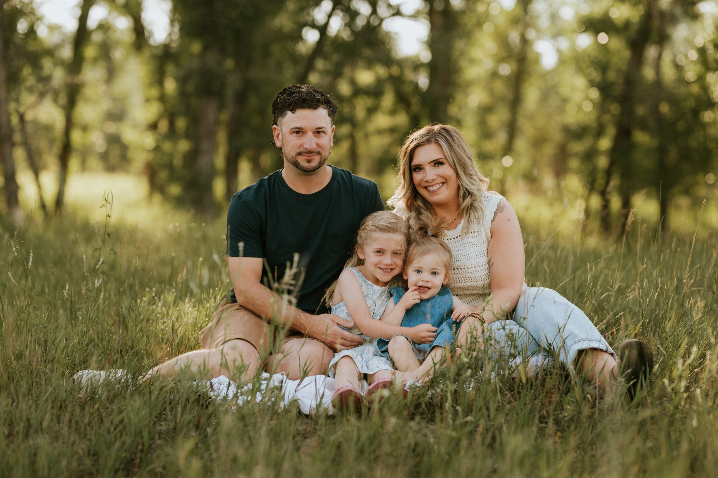 Mother and father sitting on a blanket in the green grass and tree's with their two young daughters on their lap. everyone is smiling and soaking in the sunshine of Lethbridge Alberta.