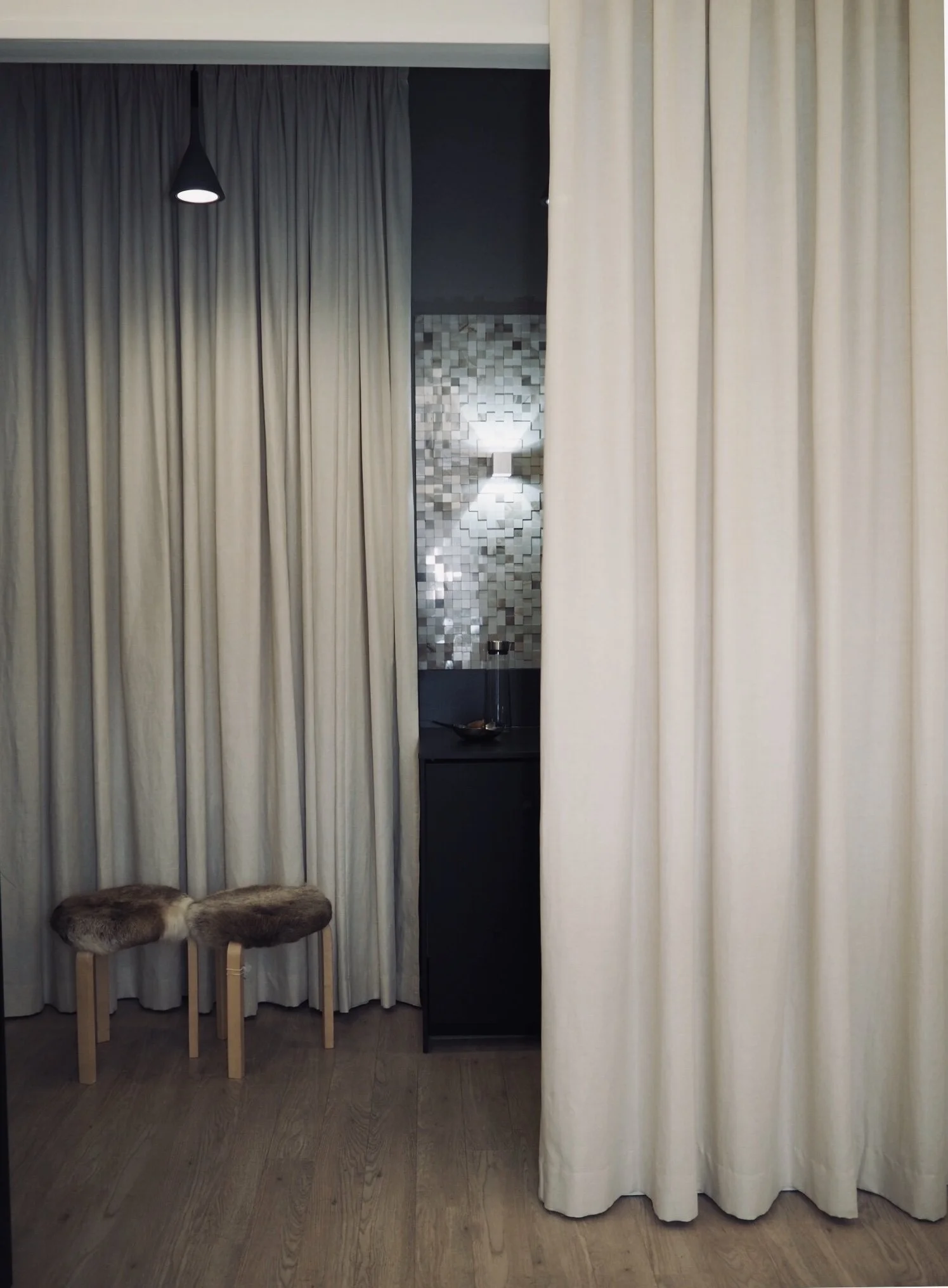 A room with closed beige curtains, a stool with fur on top, and a small black cabinet. There is a mosaic wall and a floor lamp visible behind the curtains.
