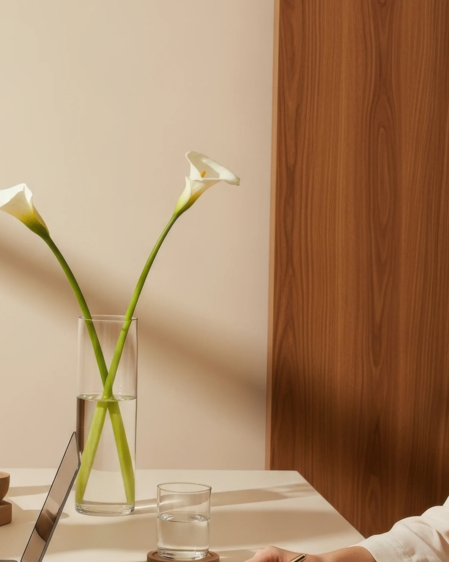 A clear glass vase holding two long-stemmed white calla lilies on a white table, with a glass of water on a coaster and a person's hand using a smartphone nearby; wooden panel and beige wall background.