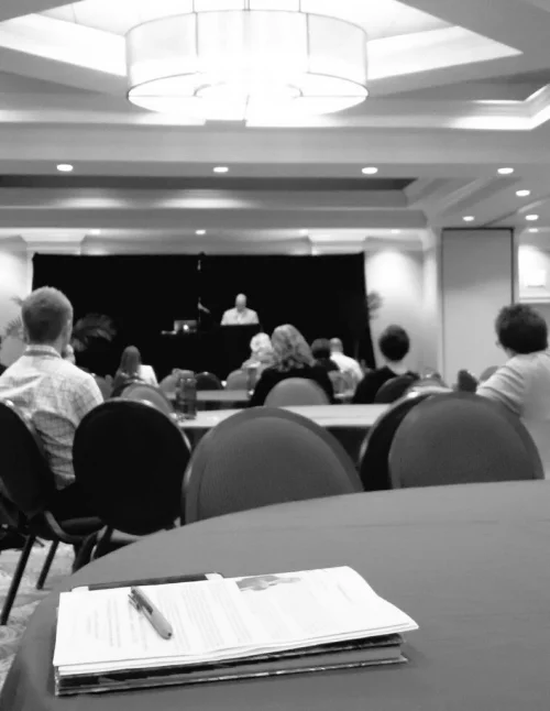 Black and white photo of a large, hotel, ballroom. In the foreground, a table with a pen and conference booklet, you can’t quite make out what it says. Beyond that are people sitting in chairs at round tables and listening to a closing keynote given by an older , male caucasian speaker at a podium.