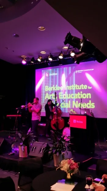 In purple-hued stage lights two musicians play. A younger black male in a short sleeve gray shirt and jeans plays trumpet with a younger white male with long hairs in a black cardigan and dark jeans plays stand-up bass. They’re both on stage with large monitors, a drum set, and key board around them a and screen bearing the conference title behind them
