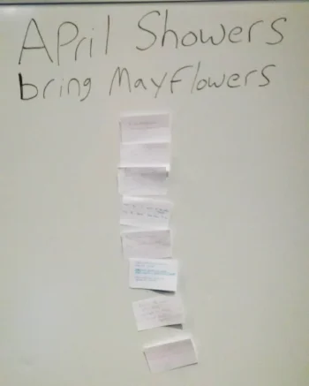 “April Showers / bring Mayflowers” is written in black lettering on a white board. Below that in a vertical column are note cards. It’s clear that there’s writing on the cards, but the writing itself is illegible in the photo.