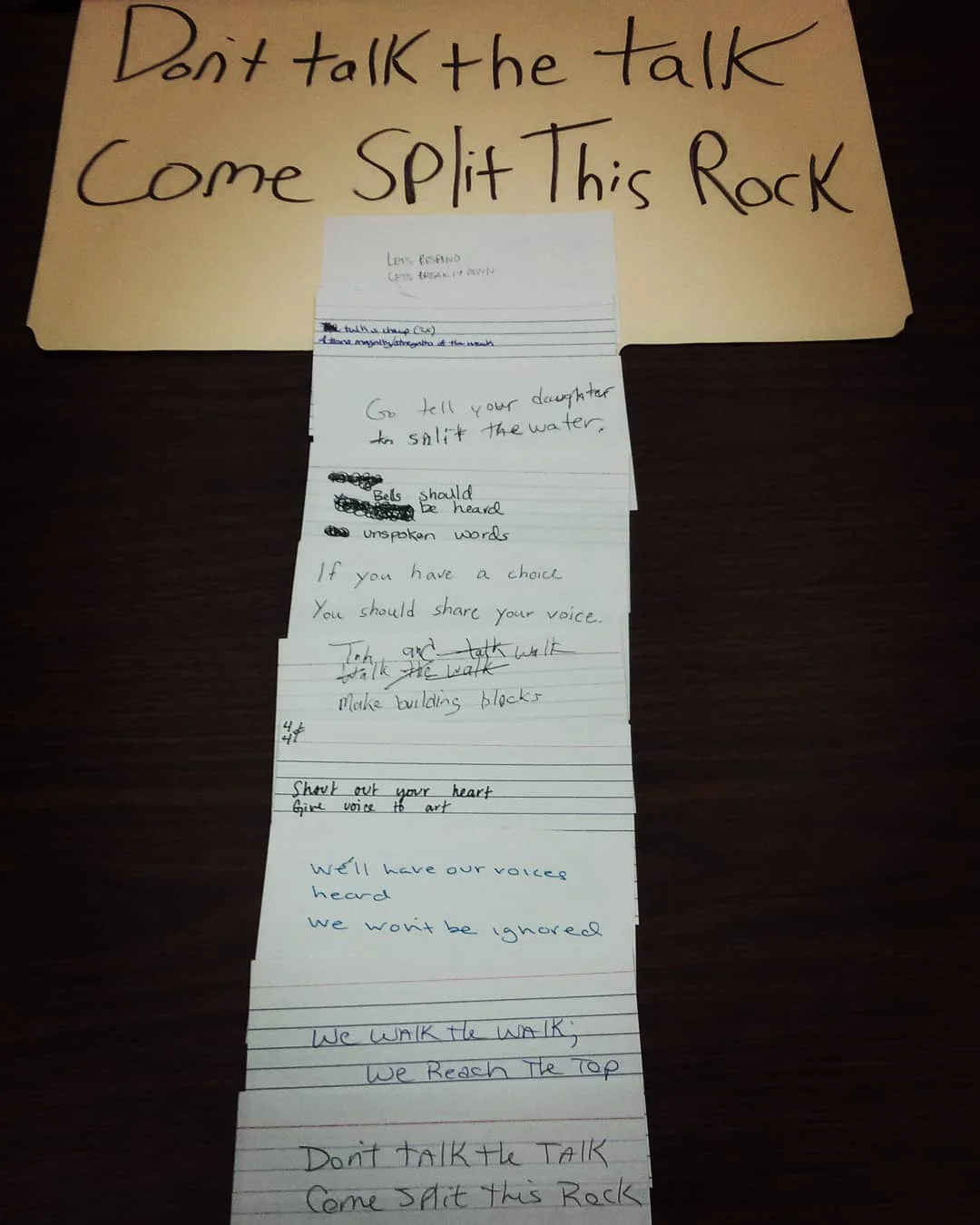 Collaborative poem made by the poets in the "Let the Words Sing" workshop at Split This Rock. At the top of a dark wooden table there’s “Don’t Talk the Talk / Come Split this Rock” written in black marker on a manila folder. Stacked vertically underneath are note cards, each with two lines of writing in pen, making up the poem.