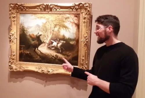 Donnie Welch, a young, white male bearded, with glasses and in a black long sleeve shirt stands in front of "The Headless Horseman Pursuing Ichabod Crane" (1858 oil on canvas) by John Quidor pointing at the art. The painting is a water color showing two horsemen, one the black, spectral, headless horseman on a black horse, reared up, the other a regular man on a white horse trying to ride away. The painting fairly small and in an ornate gold colored frame.