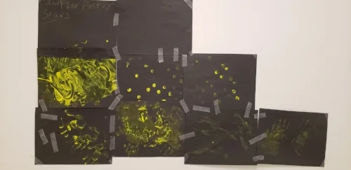 Picture of stars & constellations from a poetry workshop. Black construction paper taped together with yellow finger paint on them. To the left are two columns of three, then a column of two, then one all the way on the right. The finger painting is different in each, some dots, some swirls and some full hand prints are visible.