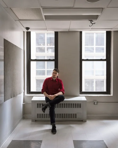 (Photo by Julianne Nash ) Donnie Welch, young caucasian male, sits on radiator by the window of a empty classroom. He’s in a red shirt, jeans, and sneakers, sitting with one leg crossed. The gray and white tile floor and white board of the classroom are visible in the foreground.