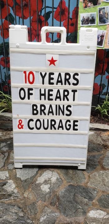 Big white letter board sign with black and red lettering. Sign has a red star at the top and reads “ 10 years / of heart / brains / & courage”. The sign is on a rocky surface with some plants and mulch from the garden visible behind it.